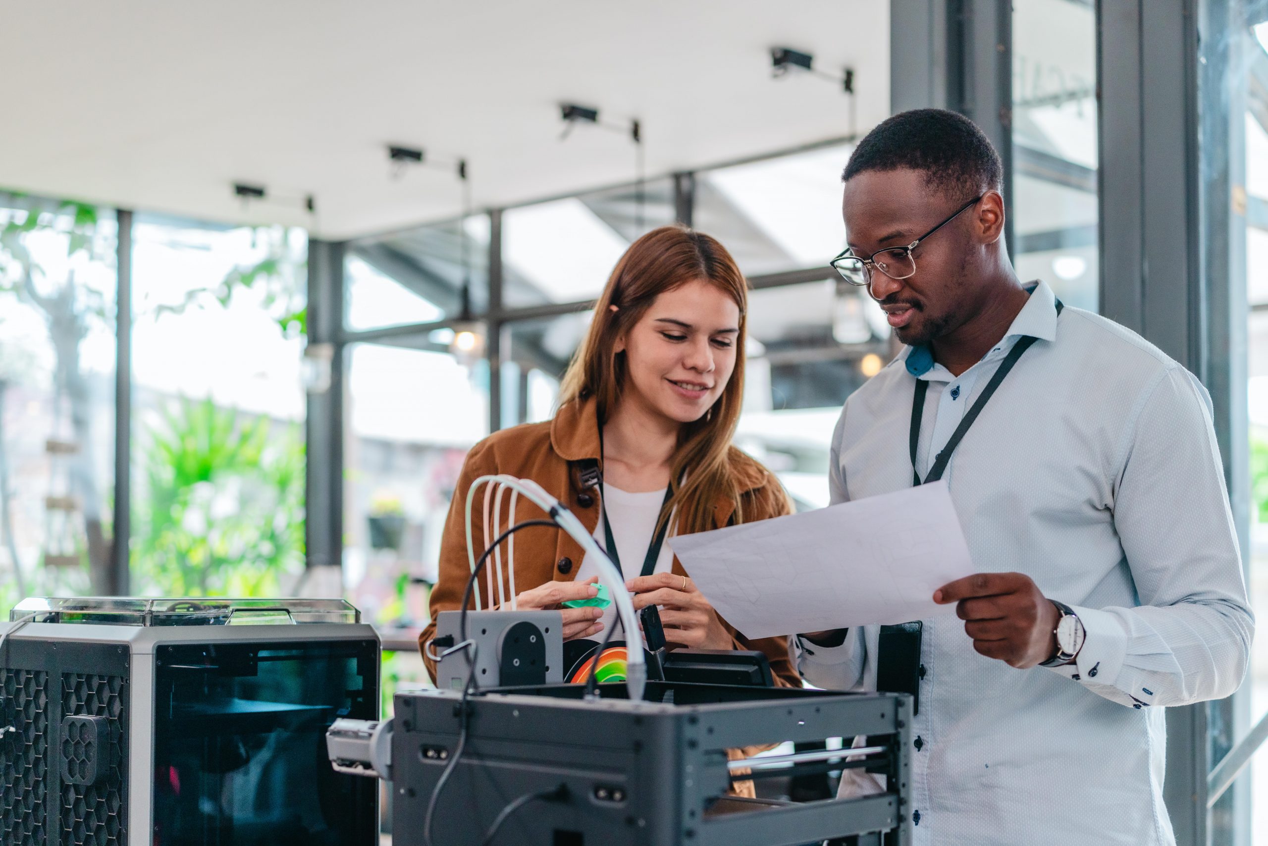 Un homme et une femme se tiennent debout près d’imprimantes 3D et examinent un plan de travail sur papier.