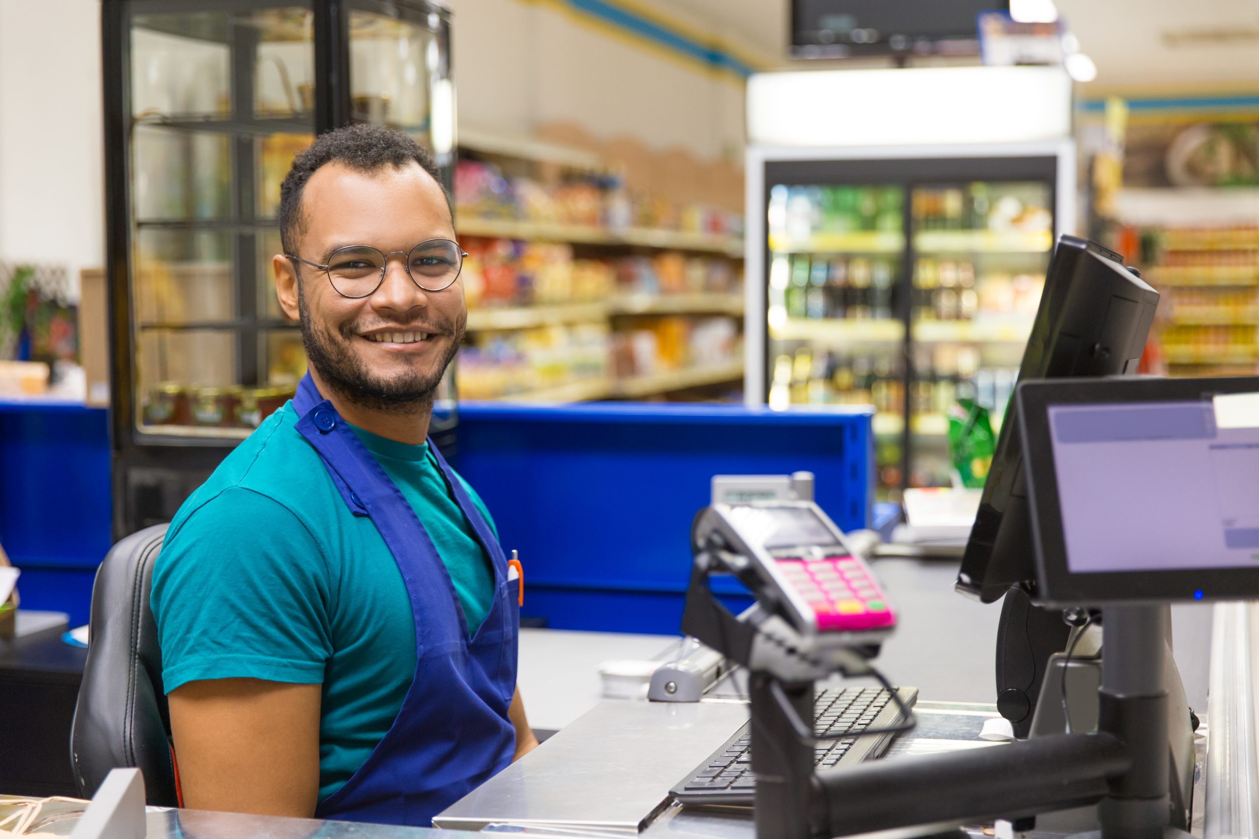 Un jeune homme souriant portant un tablier est assis derrière un écran à la caisse d’un magasin d’alimentation.